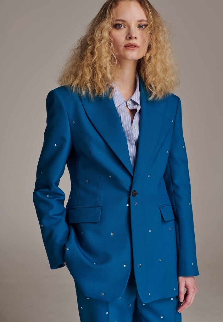 Experience flawless tailoring with this structured blazer, featuring sharp wide shoulders and a subtly cinched waist. Fully satin-lined for added luxury, it includes horn buttons and welt pockets with flaps, along with a back slit for ease of movement. Masterfully crafted in a versatile petrol, embodying precision and elegance. Adorned with striking Swarovski crystals artfully scattered across its surface. Inspired by menswear effortlessly blend sharp tailoring with a touch of glamour, ensuring a perfect fi