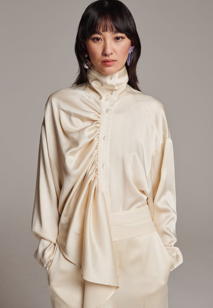 Elevate your wardrobe with this deconstructed molded top. Made in Portugal from fluid crepe satin. This gathered turtleneck top has dropped shoulders and an asymmetric dramatically gathered falling draped front panel that accentuates the loose, oversized shape. This versatile piece effortlessly transitions from day to night, making a sophisticated addition to any ensemble.