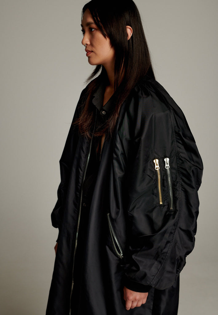 This long bomber jacket offers a perfect blend of modern style and functionality. It features two distinctive zipper closures—one in silver and the other in gold—on the sleeve, adding a bold, metallic contrast and unique detail. The elongated silhouette provides a sleek, urban look, while the durable nylon material ensures warmth and comfort.