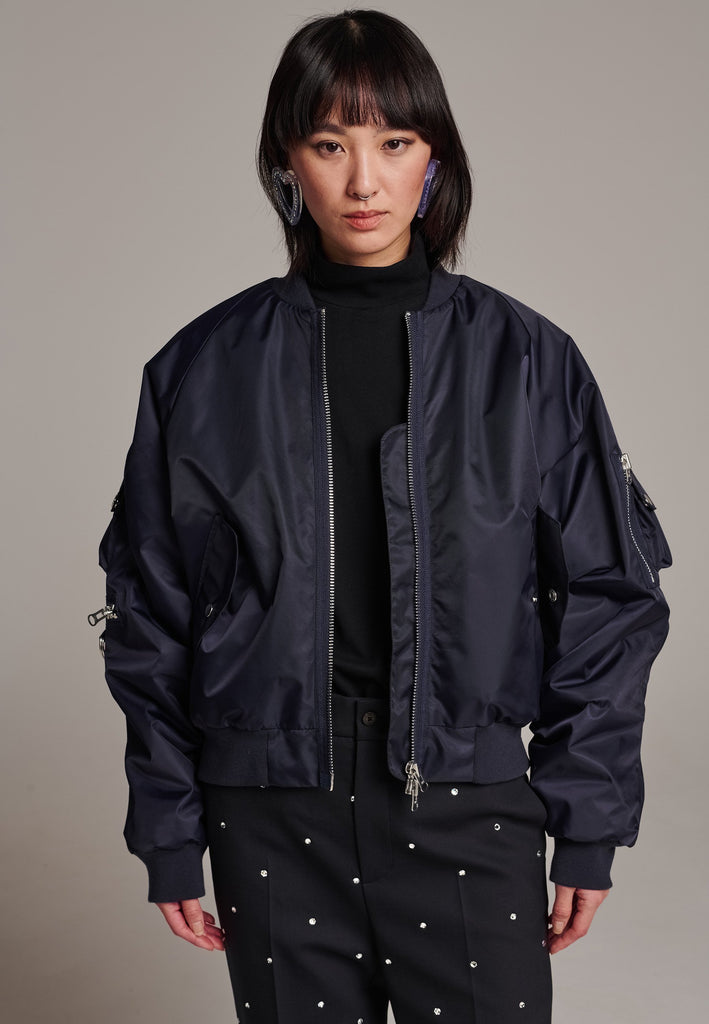 Our short bomber jacket maintains its oversized fit, crafted from crisp nylon in a deep navy blue. Adorned with silver hardware and zippers, this jacket adds a touch of edge to any outfit. Whether styled with tailored pants or a long skirt, it effortlessly enhances your look with a cool, contemporary flair.