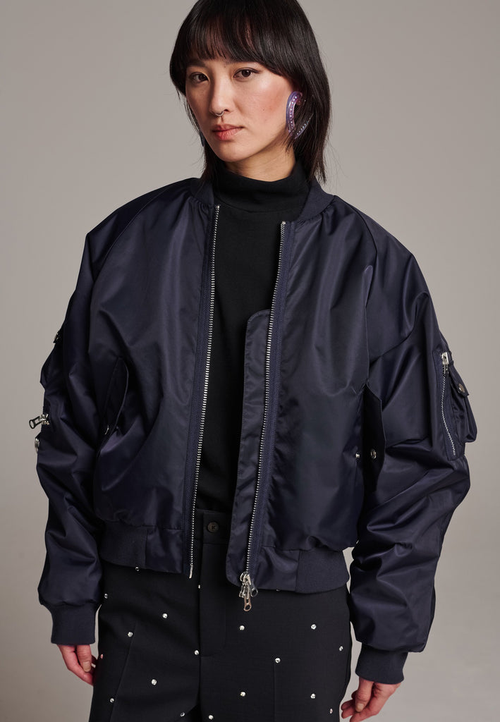 Our short bomber jacket maintains its oversized fit, crafted from crisp nylon in a deep navy blue. Adorned with silver hardware and zippers, this jacket adds a touch of edge to any outfit. Whether styled with tailored pants or a long skirt, it effortlessly enhances your look with a cool, contemporary flair.