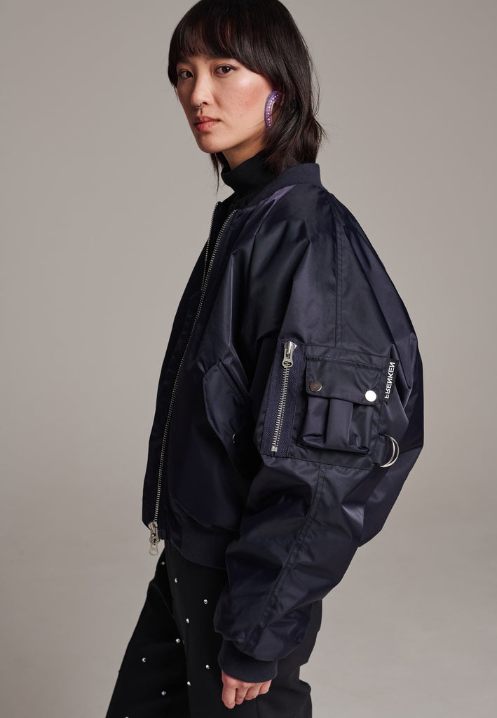 Our short bomber jacket maintains its oversized fit, crafted from crisp nylon in a deep navy blue. Adorned with silver hardware and zippers, this jacket adds a touch of edge to any outfit. Whether styled with tailored pants or a long skirt, it effortlessly enhances your look with a cool, contemporary flair.