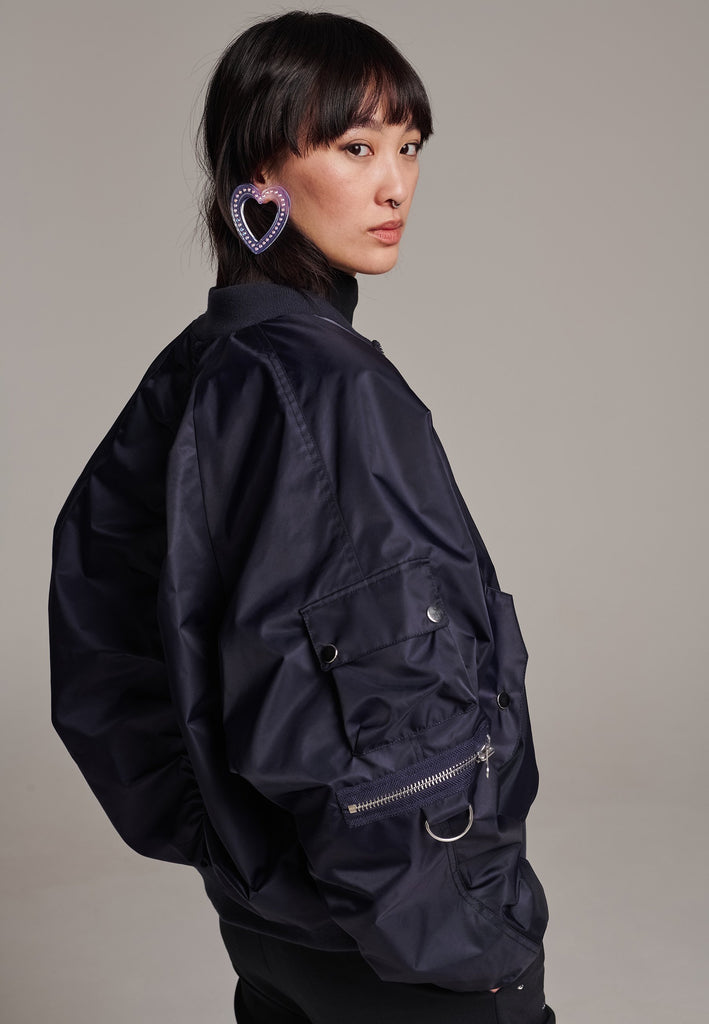 Our short bomber jacket maintains its oversized fit, crafted from crisp nylon in a deep navy blue. Adorned with silver hardware and zippers, this jacket adds a touch of edge to any outfit. Whether styled with tailored pants or a long skirt, it effortlessly enhances your look with a cool, contemporary flair.