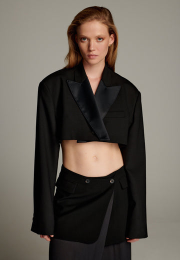 This short blazer is a contemporary take on classic tailoring, featuring a cropped length and a sleek silhouette. It is crafted from a black fabric with a satin-finish lapel, adding a touch of sophistication and contrast to the design. The blazer has long, structured sleeves with clean lines and a minimalist aesthetic, exuding modern elegance. Perfect for pairing with high-waisted trousers or skirts, it combines boldness with versatility, making it a statement piece for fashion-forward ensembles.