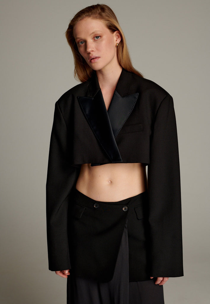 This short blazer is a contemporary take on classic tailoring, featuring a cropped length and a sleek silhouette. It is crafted from a black fabric with a satin-finish lapel, adding a touch of sophistication and contrast to the design. The blazer has long, structured sleeves with clean lines and a minimalist aesthetic, exuding modern elegance. Perfect for pairing with high-waisted trousers or skirts, it combines boldness with versatility, making it a statement piece for fashion-forward ensembles.