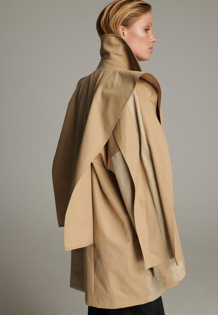 Inspired by a men's coat, FRENKEN offers a refined, contemporary twist on classic outerwear. Designed with dual-tone camel panels that create a subtle yet striking contrast, it exudes effortless sophistication. Featuring squared draped fabric, the coat showcases a modern, architectural design. Made from water-resistant cotton, it combines style, versatility, and functionality for everyday wear.